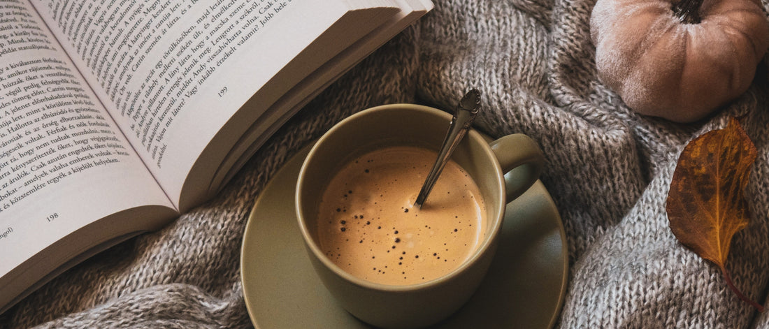 A cup of hot coffee and an open book on a knitted grey blanket. 