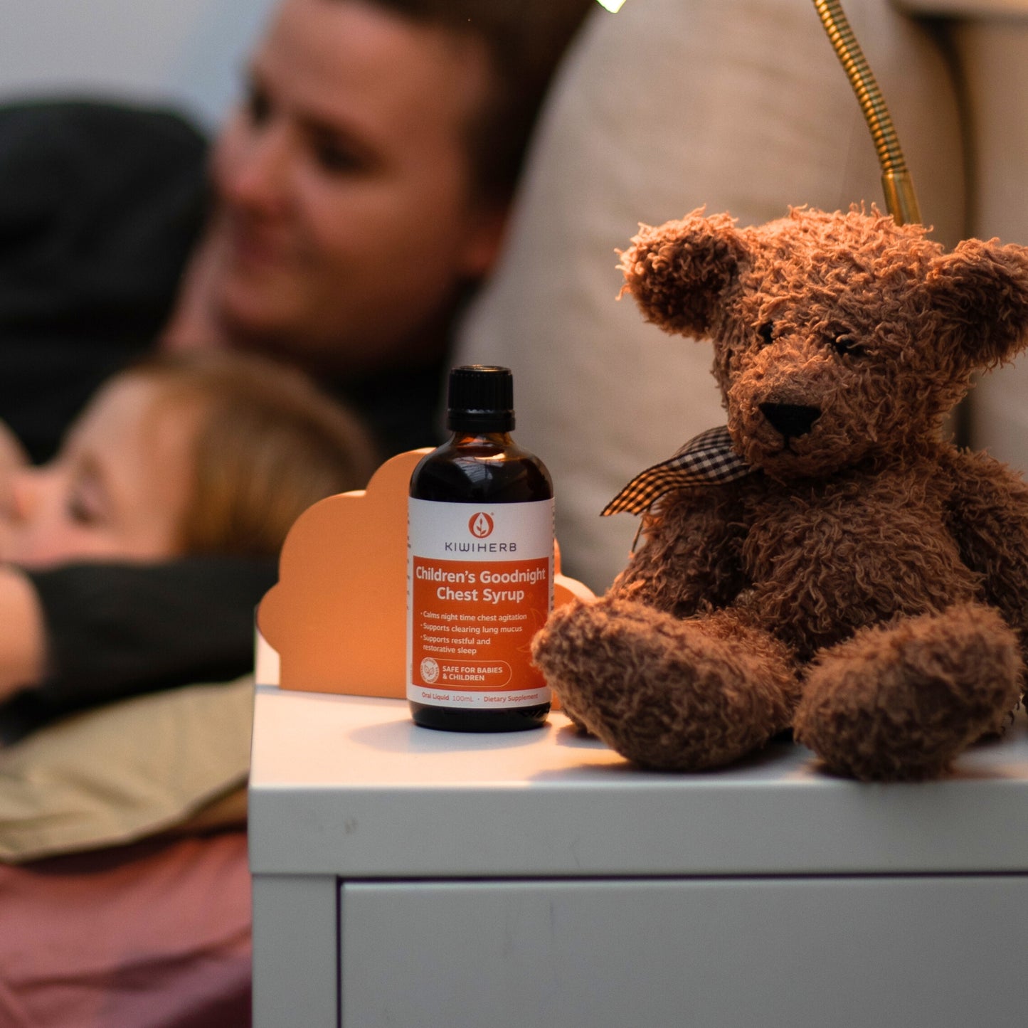 A bottle of Children's Goodnight Chest Syrup near a brown teddy bear on a children's bedside table.