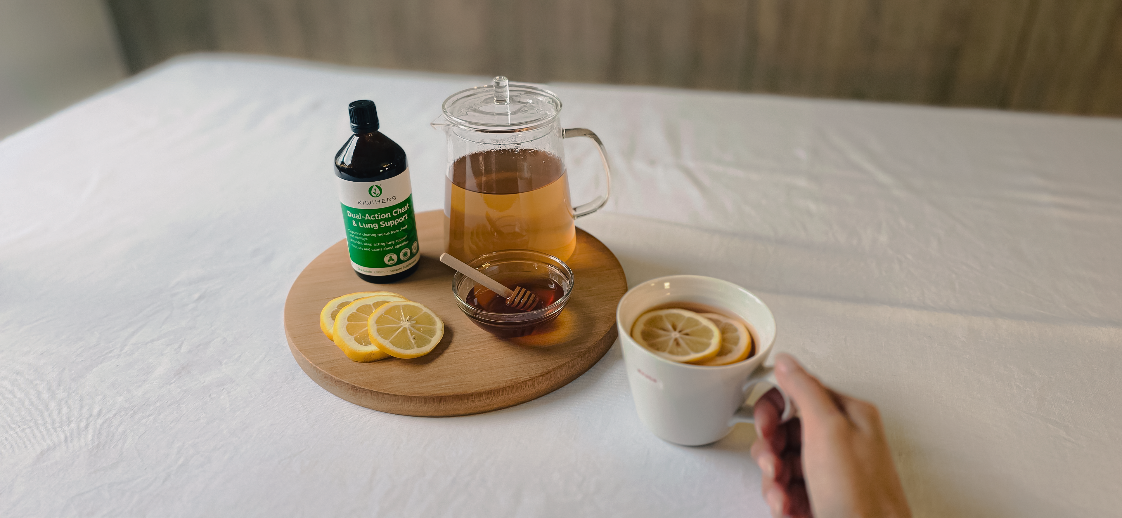 Tea-making setup with lemon, honey, and a bottle of  Dual Action Chest And Lung Support  on a wooden board, with a hand holding a cup of tea.