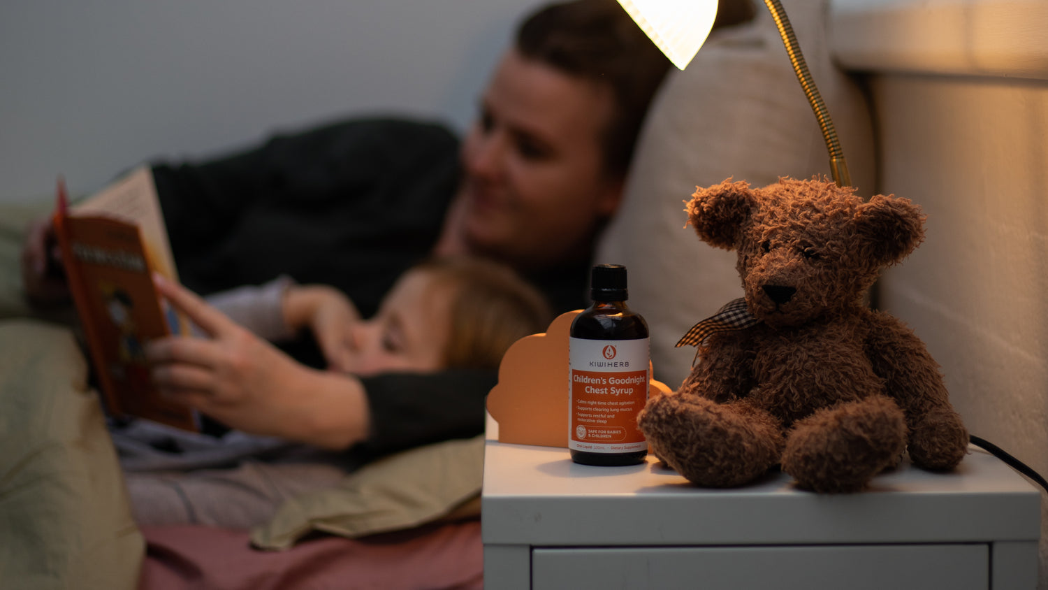 Person lying on a couch reading a book with a teddy bear and bottle of nighttime cough syrup on a table in the foreground.